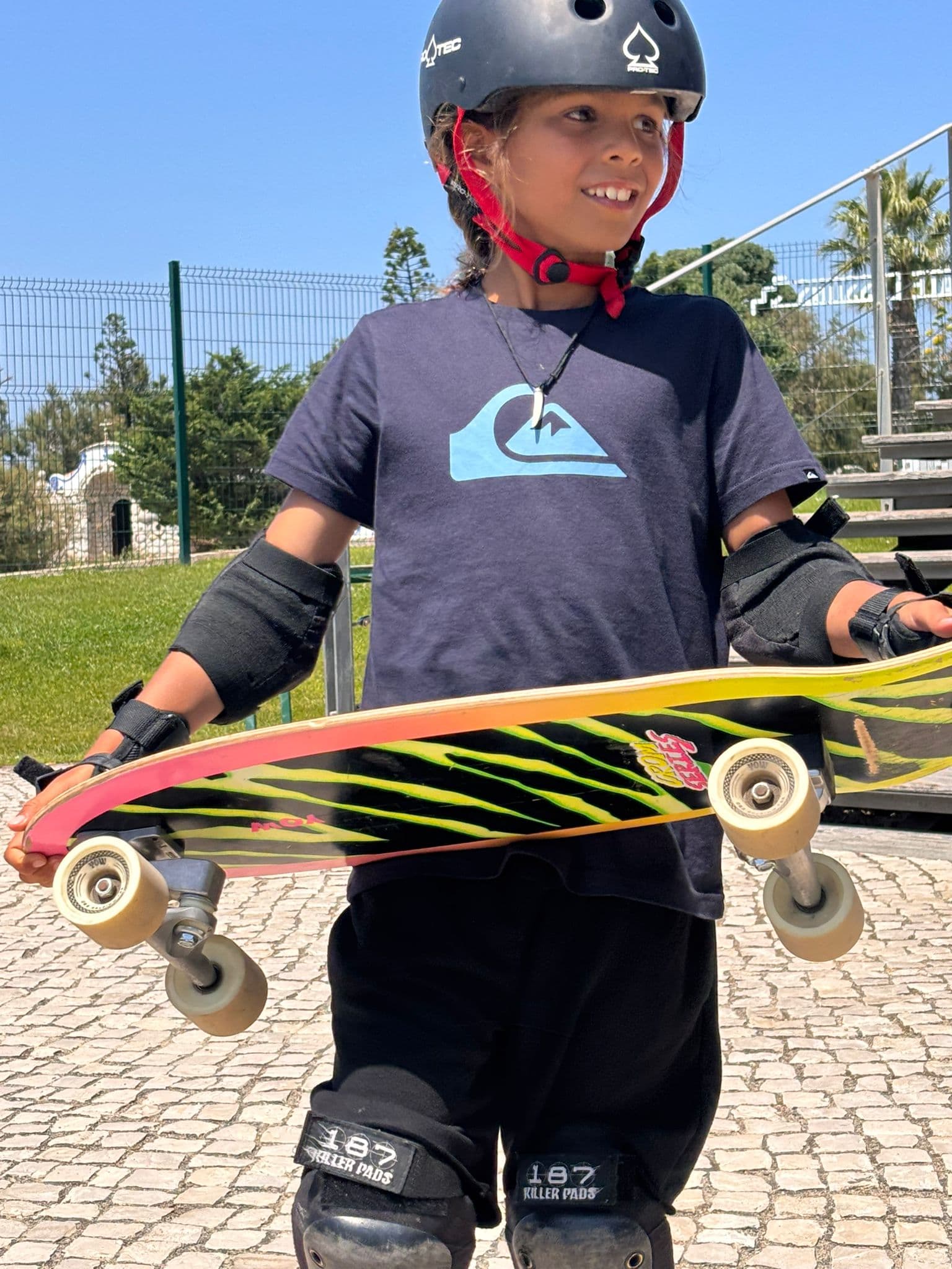 Un garçon avec des protections de skate sourit en tenant un longboard coloré